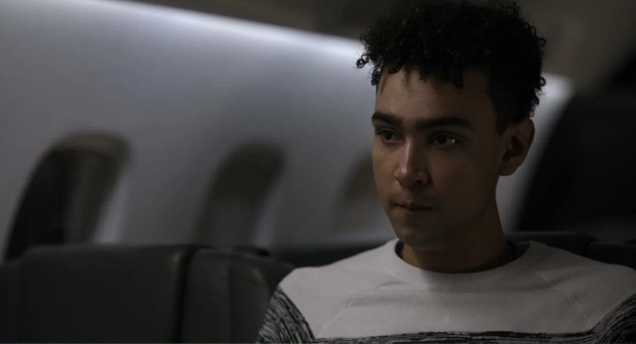 Gran Turismo (2023) movie still: medium — A passenger sits in a dark, out-of-focus airplane cabin. The man stares forward with a neutral expression…