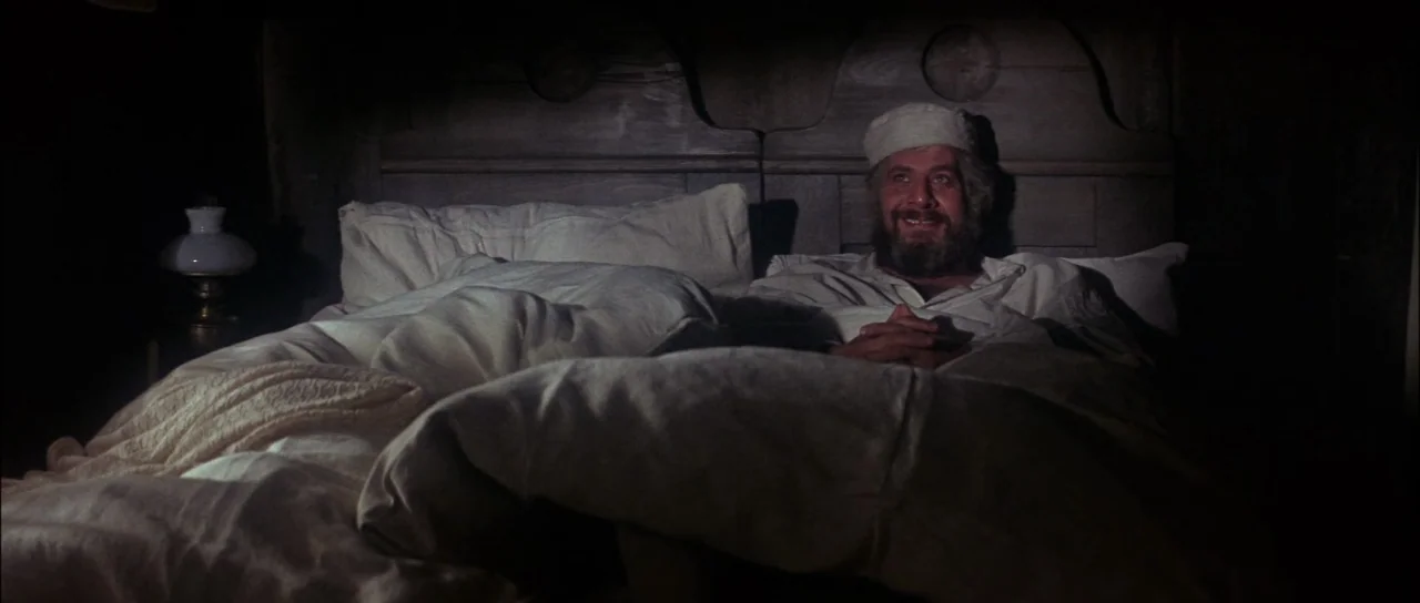 Fiddler On The Roof (1971) movie still: medium — A man lies in a bed with a wooden headboard in a dark room. He smiles while resting his hands on…