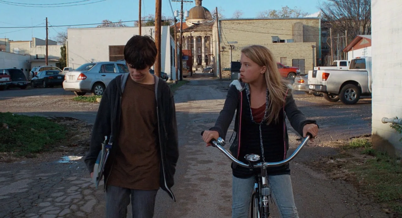 Boyhood (2014) movie still: fullbody — An alleyway between small town buildings leads toward a domed courthouse in the distance. A teenage boy walks…