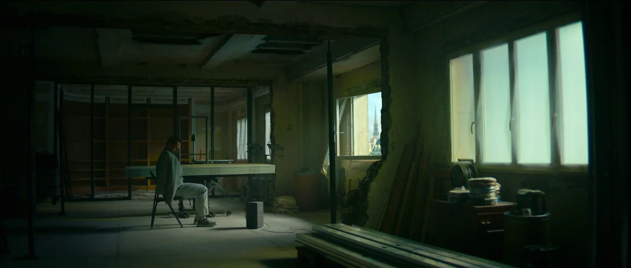 The Killer (2023) movie still: wide — A spacious, unfinished room features exposed wall frames and construction debris on the floor. A man sits…
