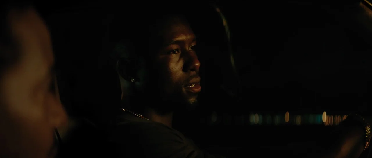 Moonlight (2016) movie still: medium — A dark car interior at night with a man driving. The driver turns his head slightly to look at a passenger in…