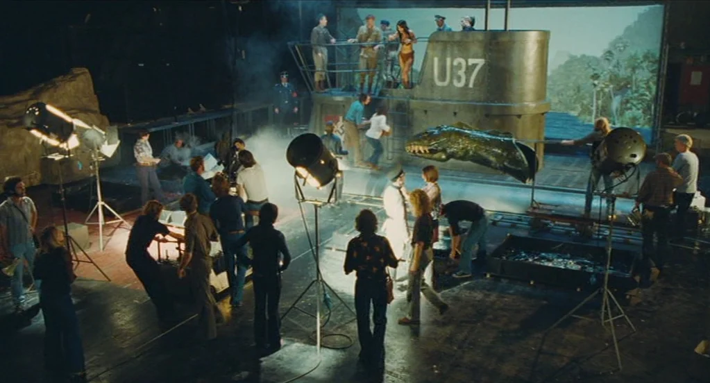 Something in the Air (2012) movie still: wide — A crowded, dimly lit film soundstage contains a large submarine prop marked U37. Crew members walk…