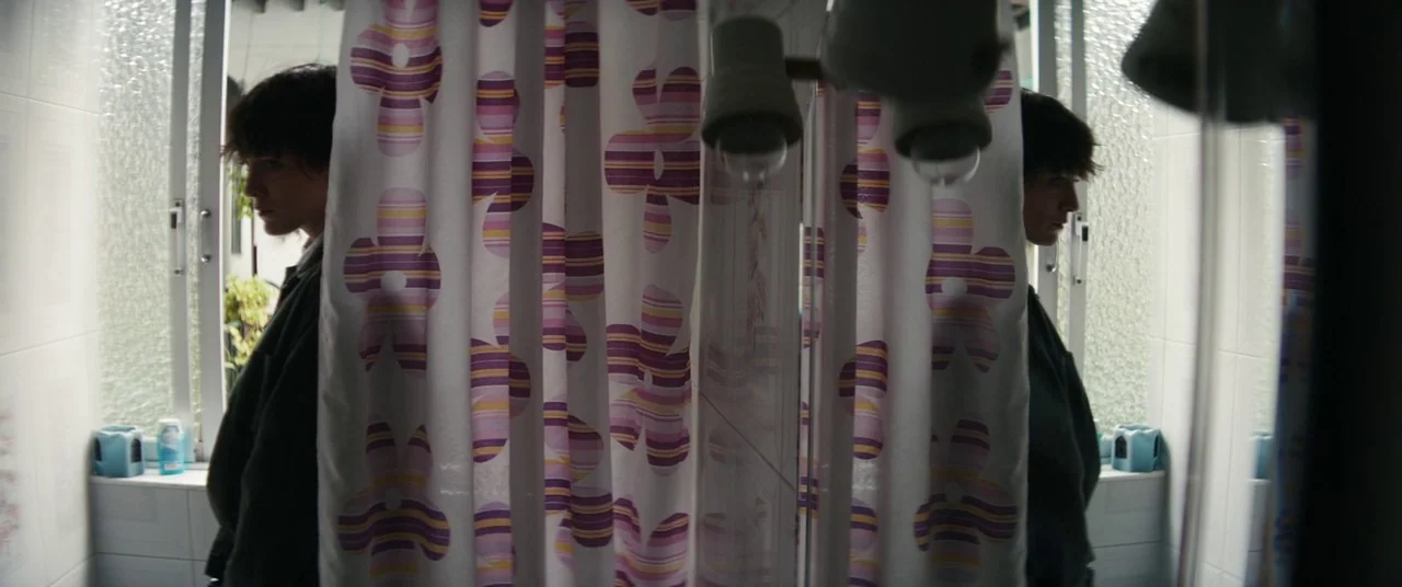 The Rhythm Section (2020) movie still: medium — A bathroom setting features a white tiled area with a window and mirror. The subject stands before…