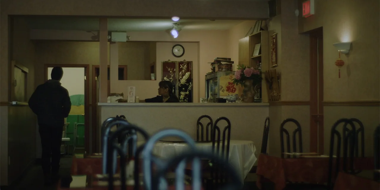 Lobos (2019) movie still: medium — The interior of an Asian restaurant with round tables and metal frame chairs. A man walks toward the service…