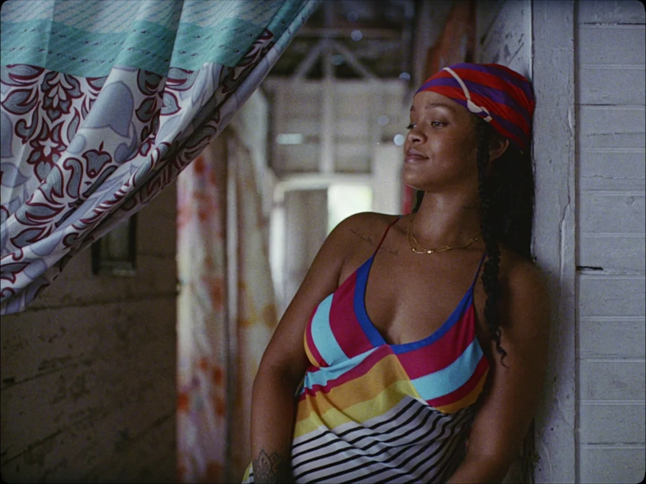 Guava Island (2019) movie still: medium — A wooden porch area features floral curtains and white painted plank walls. A woman leans against the wall…
