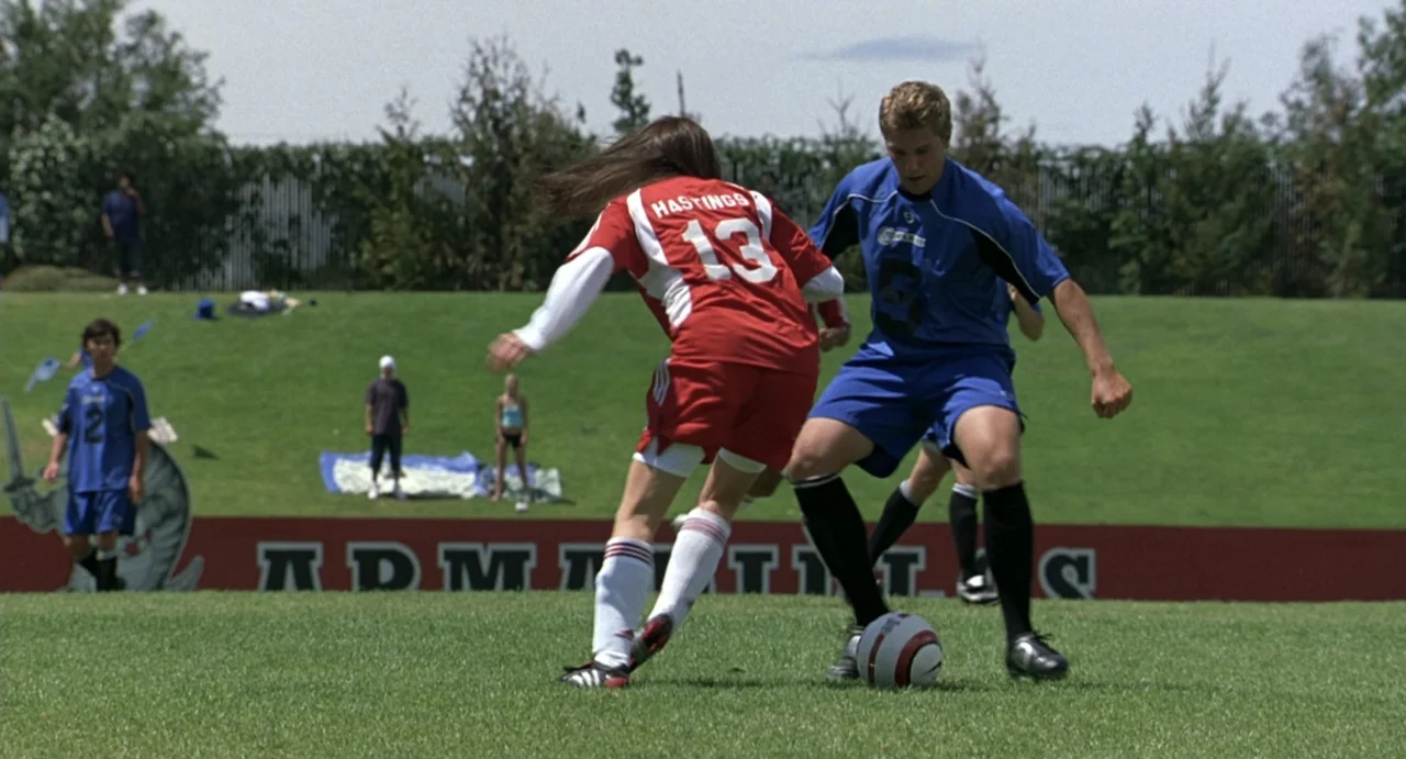 She's the Man (2006) movie still: fullbody — Two soccer players compete for control of the ball on a green grass pitch. A red barrier with white…