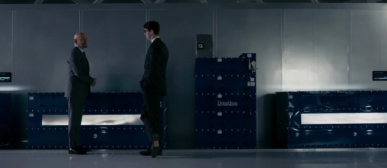 Self/less (2015) movie still: wide — A clean laboratory room features metallic walls and rows of large, blue industrial equipment. Two men in dark…