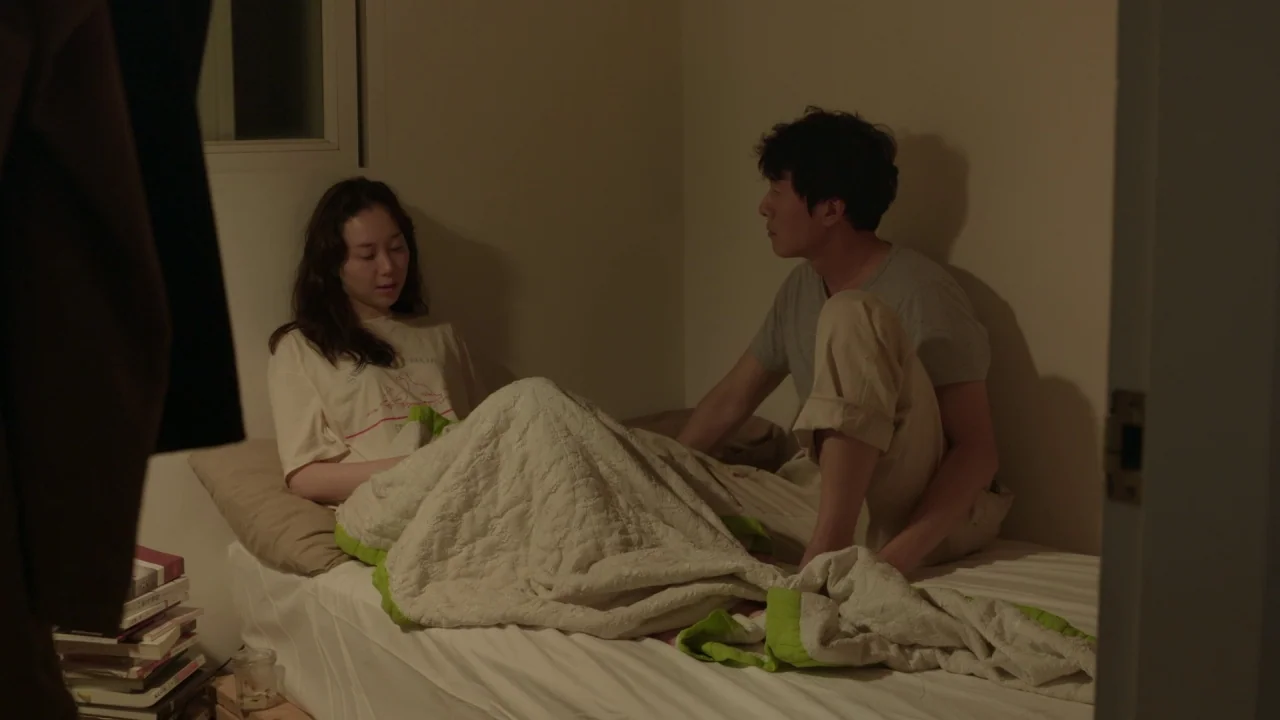 Yourself and Yours (2016) movie still: medium — A small room features a bed pushed against a corner with neutral-colored walls. A young man and…