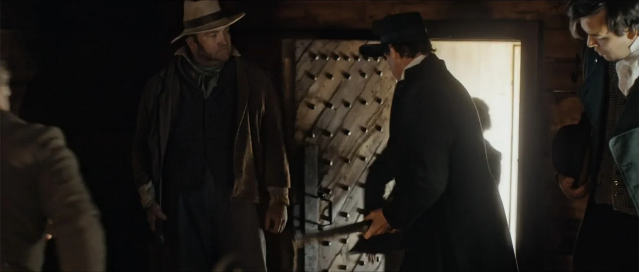 Out of Liberty (2019) movie still: medium — The interior of a rustic log cabin features a heavy studded wooden door. Three men in mid-19th-century…