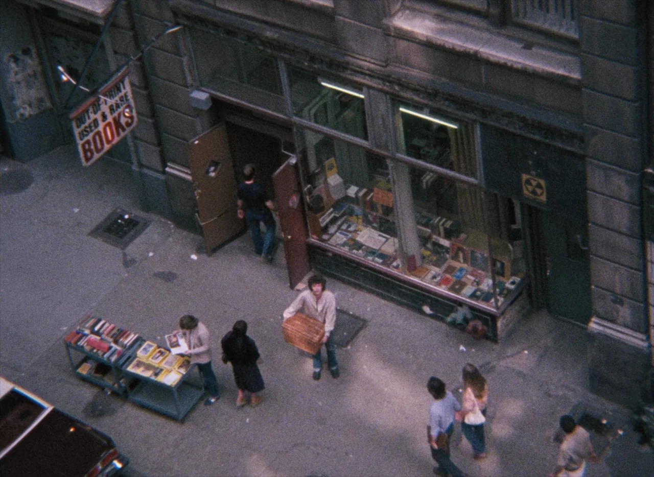 Basket Case (1982) movie still: wide — A sidewalk in front of a storefront features book carts and pedestrians. A man walks toward the entrance…