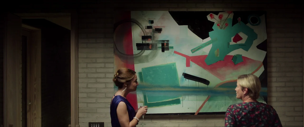The Feast (2021) movie still: medium — A painted brick wall displays a large abstract painting with geometric shapes and brushstrokes. Two women…