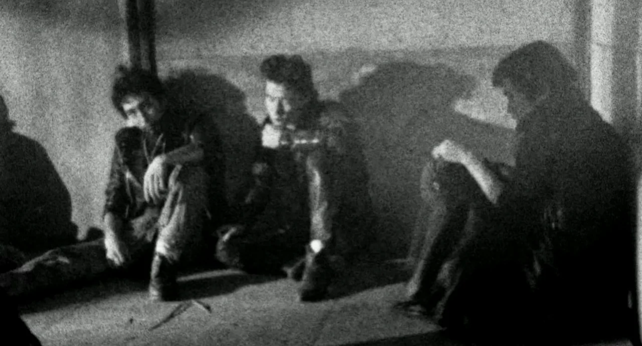 Burst City (1982) movie still: medium — A dimly lit room with a concrete floor and plain textured wall behind. Three young men sit in a row against…