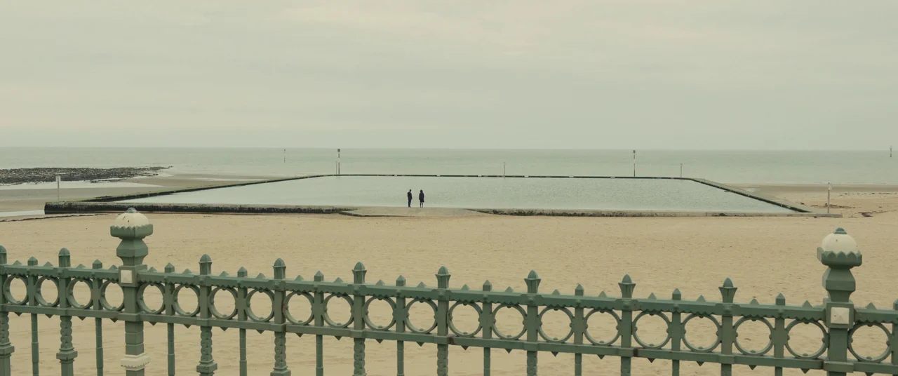 Empire of Light (2022) movie still: wide — A wide coastal area features a large rectangular tidal pool on a sandy beach. Two people stand together…