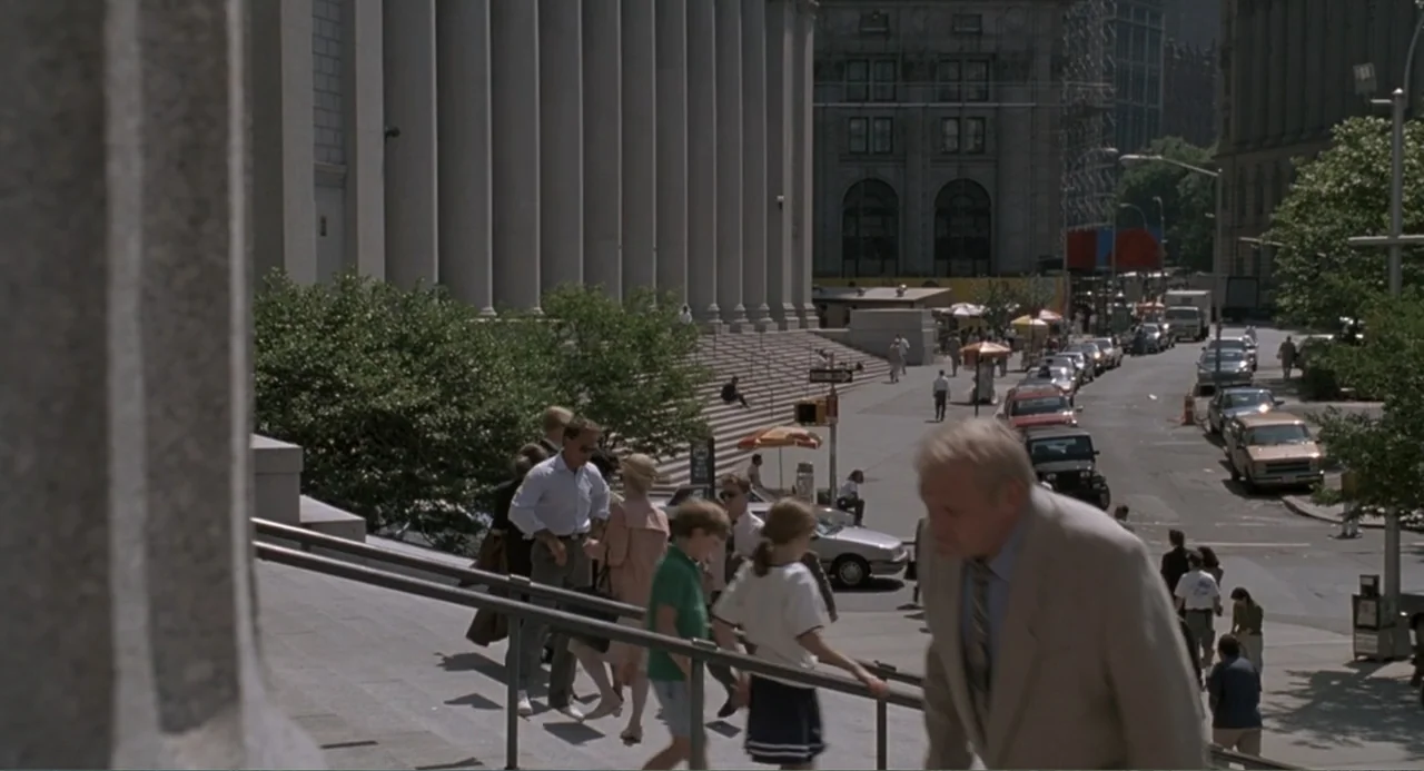F/X2 (1991) movie still: wide — A stone building with large columns stands above a busy urban sidewalk area. Several pedestrians walk down the…