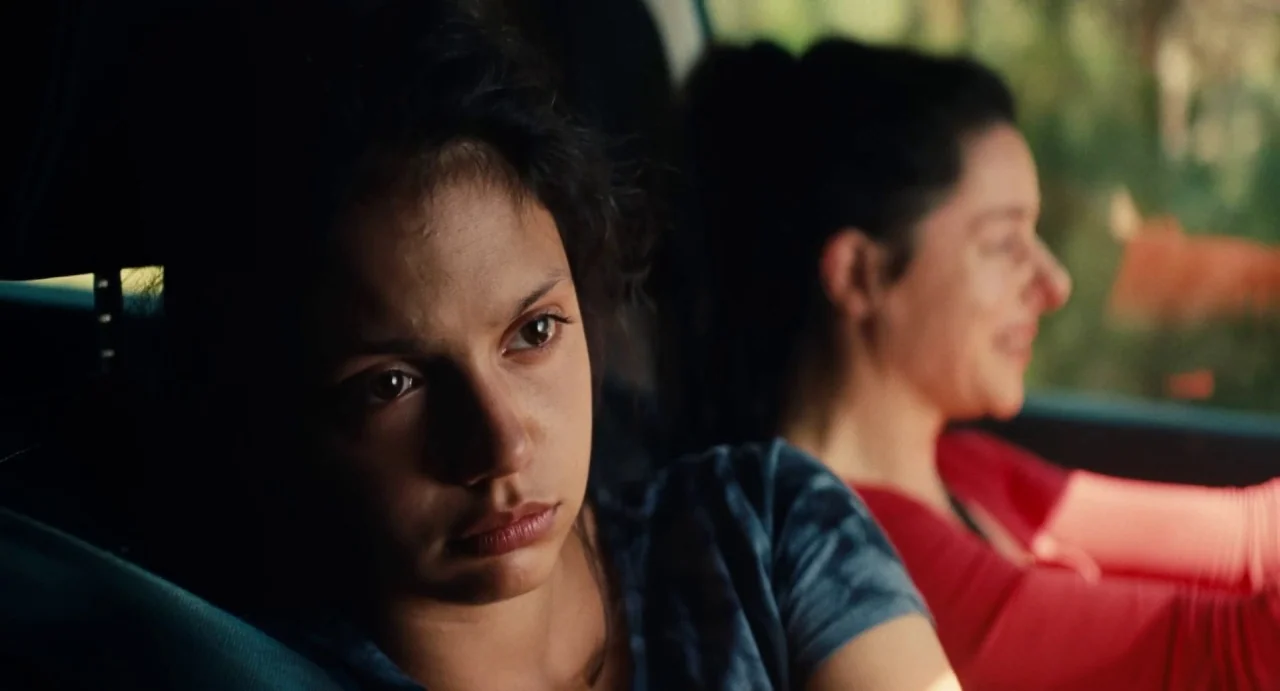 Ava (2017) movie still: closeup — Inside a moving vehicle, a young woman in the backseat looks out the window. Beside her, a driver with her hair…