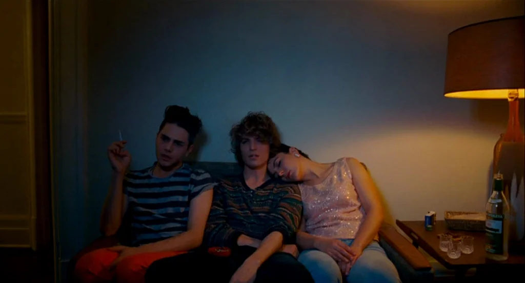 Heartbeats (Les Amours Imaginaires) (2010) movie still: medium — Three people sit on a couch inside a dark room with minimal furniture. One man…