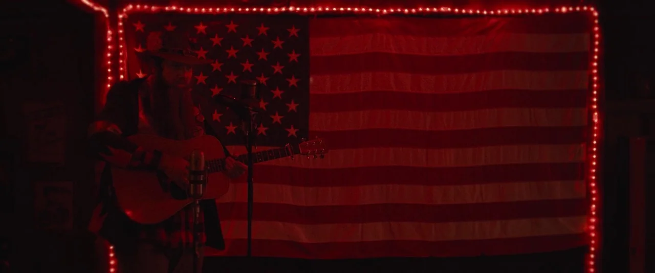 Blaze (2018) movie still: medium — A dark space features a large American flag illuminated by red string lights. A bearded man plays an acoustic…