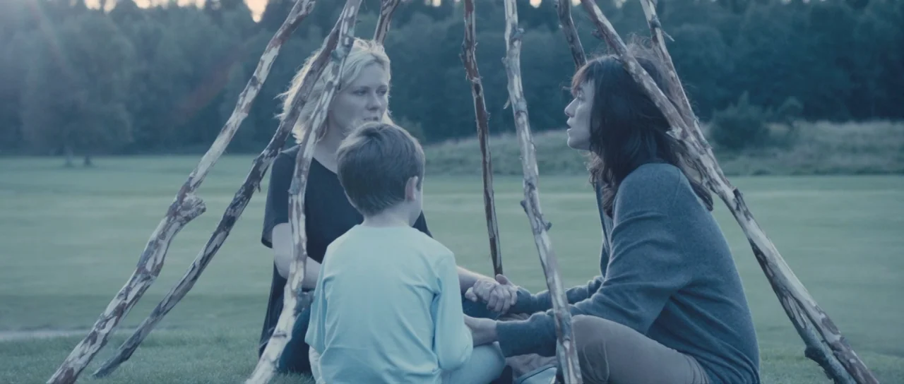 Melancholia (2011) movie still: medium — Two women and a young boy sit in a field within a teepee-like structure. They hold hands while positioned…