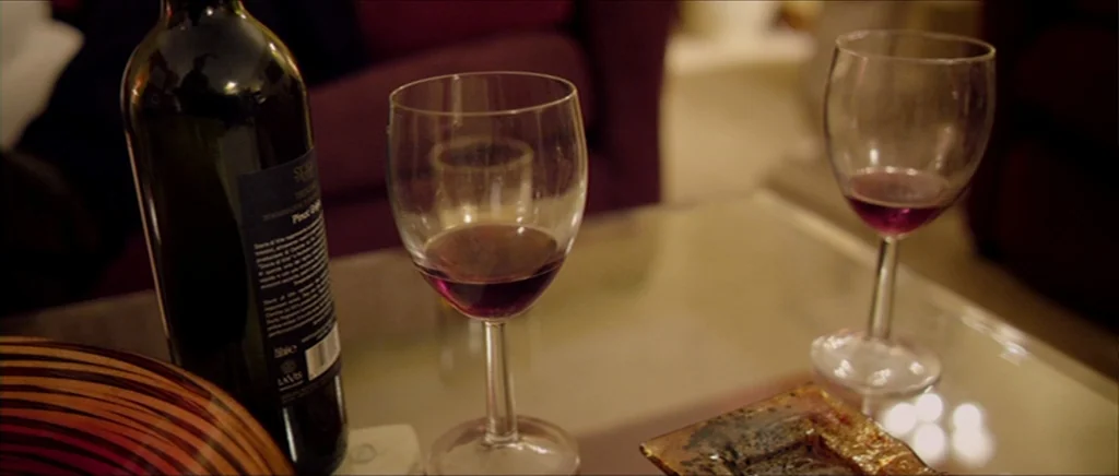 Kill List movie still: closeup — A glass coffee table holds a bottle of red wine and two stemmed glasses. The blurred background shows parts of a…