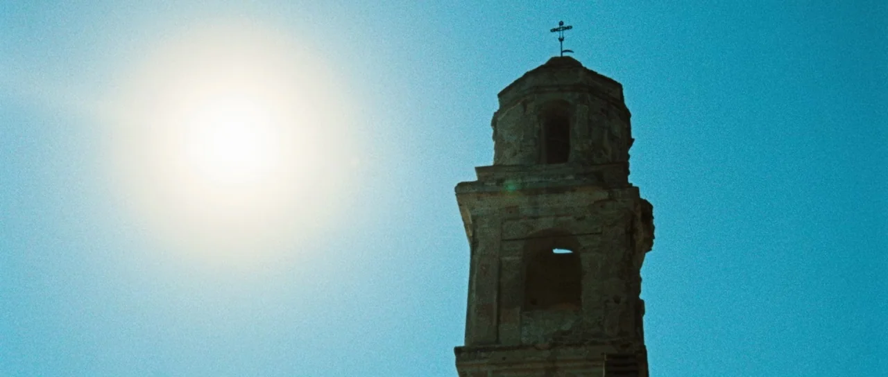 Let The Corpses Tan (2017) movie still: establishing — A stone bell tower with a small cross on top stands against a sky. The sun is visible as a…