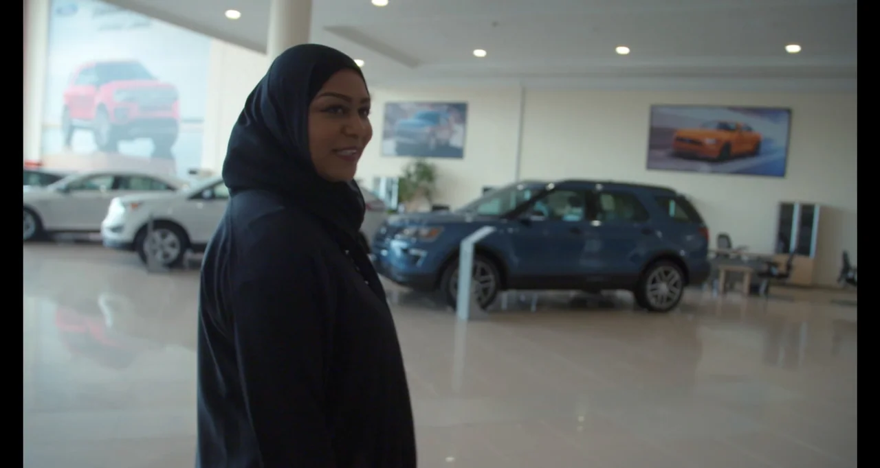 Saudi Women's Driving School (2019) movie still: medium — A brightly lit car showroom with polished floors and several modern vehicles parked…