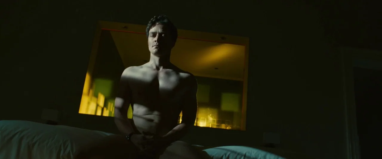 Trance (2013) movie still: medium — A dark hotel room with a glowing yellow window panel behind the central subject. A man sits shirtless on the…