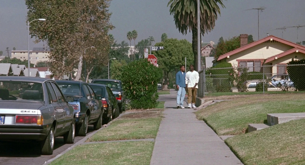Boyz In The Hood (1991) movie still: wide — A residential street lined with palm trees and parked cars features houses on either side. Two young men…