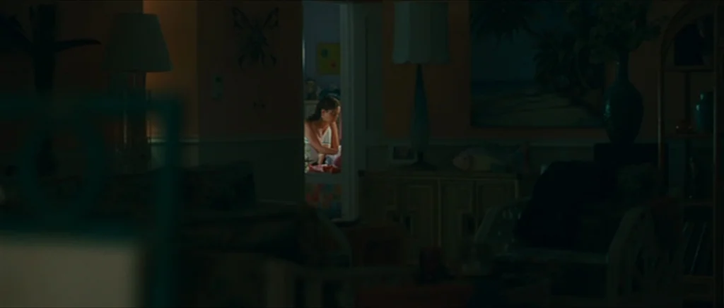 Away We Go (2009) movie still: wide — A dimly lit living room with rattan furniture and wall art sits in shadow. A young woman is visible through a…