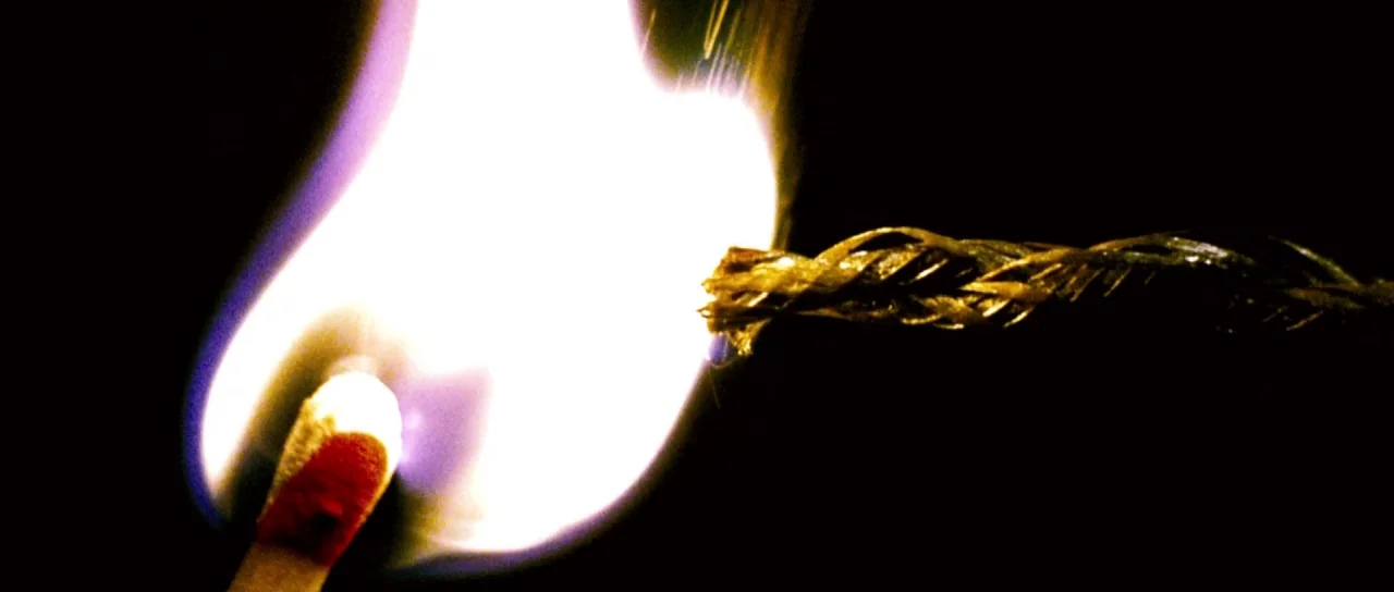 Down With Love (2003) movie still: closeup — A match flame burns brightly against a black void. The flame tip touches the end of a braided fiber rope.