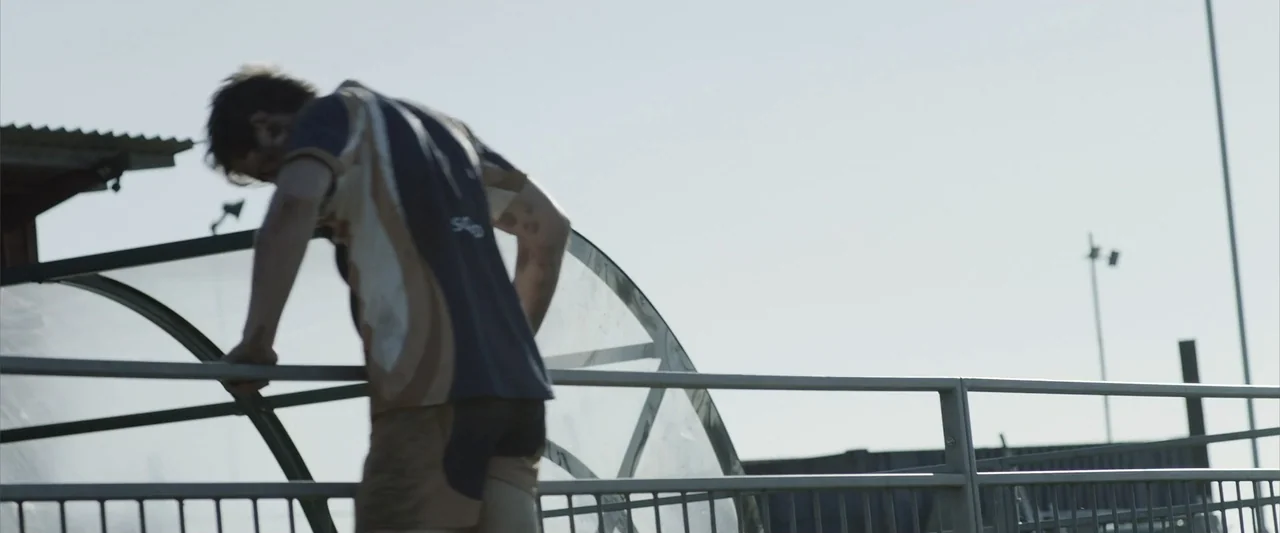 The Thorn On His Side (2014) movie still: medium — An outdoor sports facility with a curved dugout structure under a clear sky. A young man in…