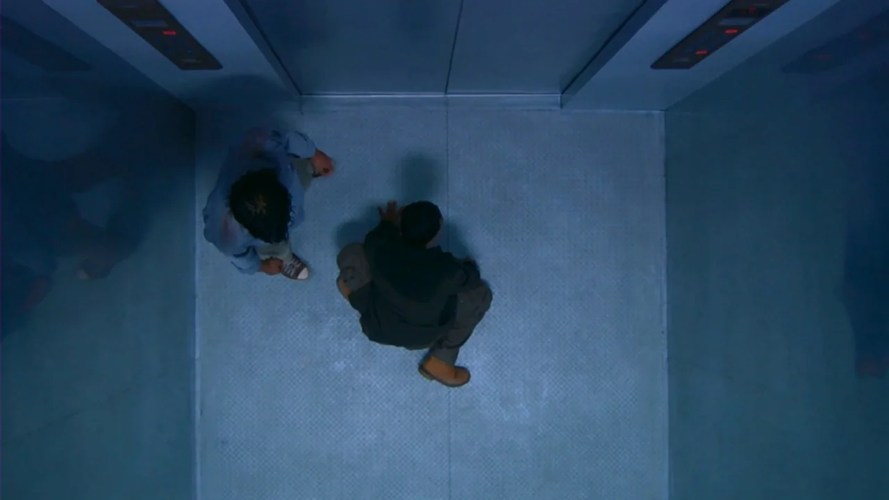 Merantau (2009) movie still: wide — A modern metal elevator car with plain panels and a textured diamond-plate floor surface. Two men occupy the…