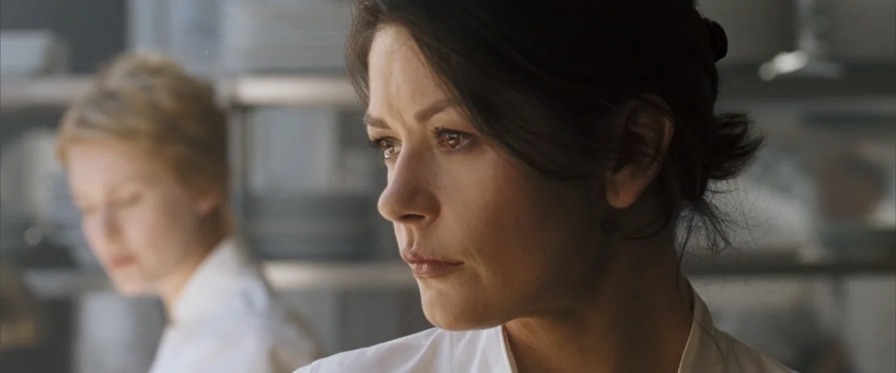No Reservations (2007) movie still: closeup — A professional kitchen features stainless steel shelving and blurry background prep areas. A woman in…