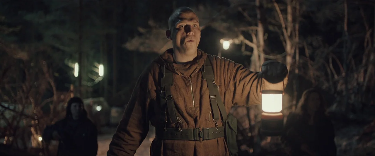 Azrael (2024) movie still: medium — A dark, wooded environment with blurred trees and faint lights in the distant background. A bald man wearing a…