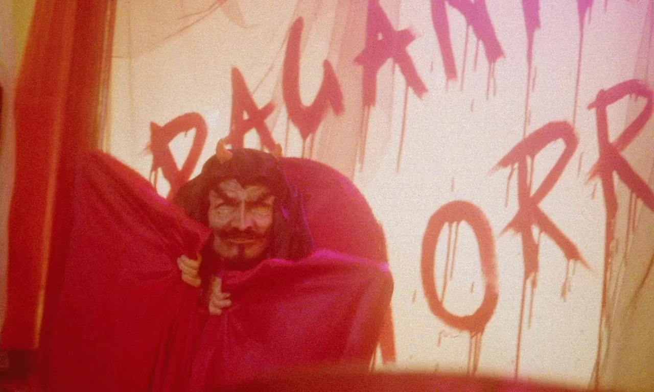 Paganini Horror (1989) movie still: medium — A figure in a red cape stands before a wall with red painted text. The text appears to read like a…