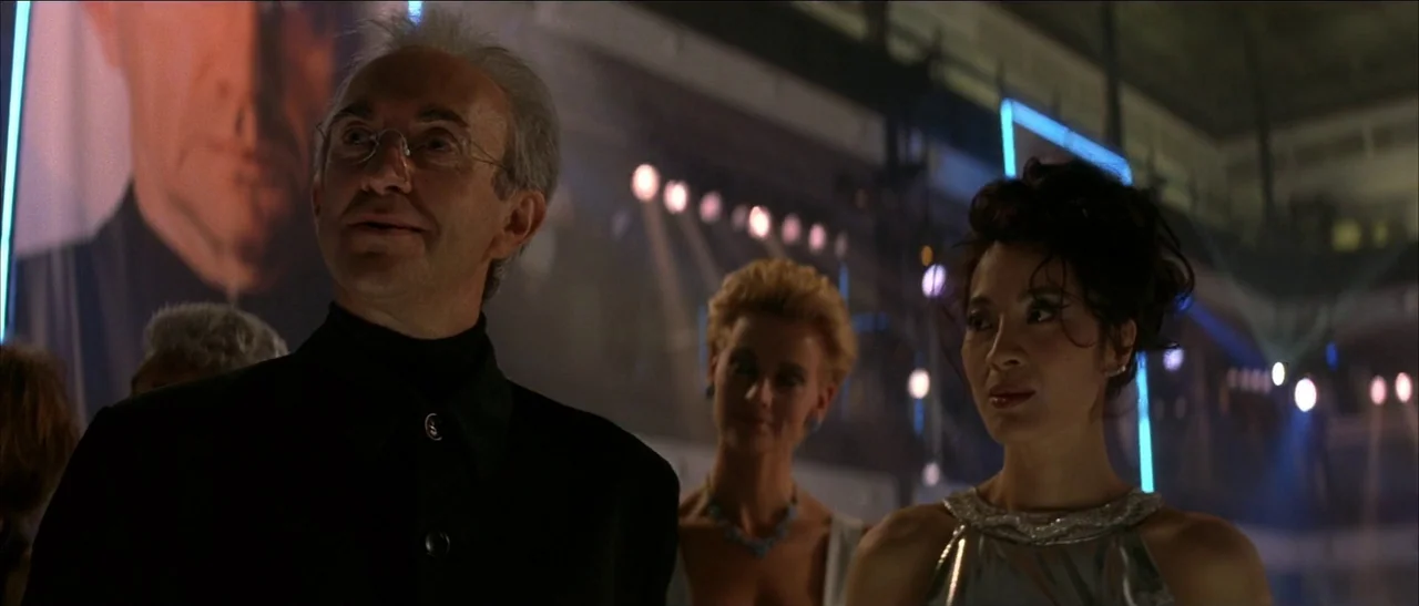Tomorrow Never Dies (1997) movie still: medium — A dimly lit indoor gala event with multiple figures in formal wear. A man and a woman stand in the…