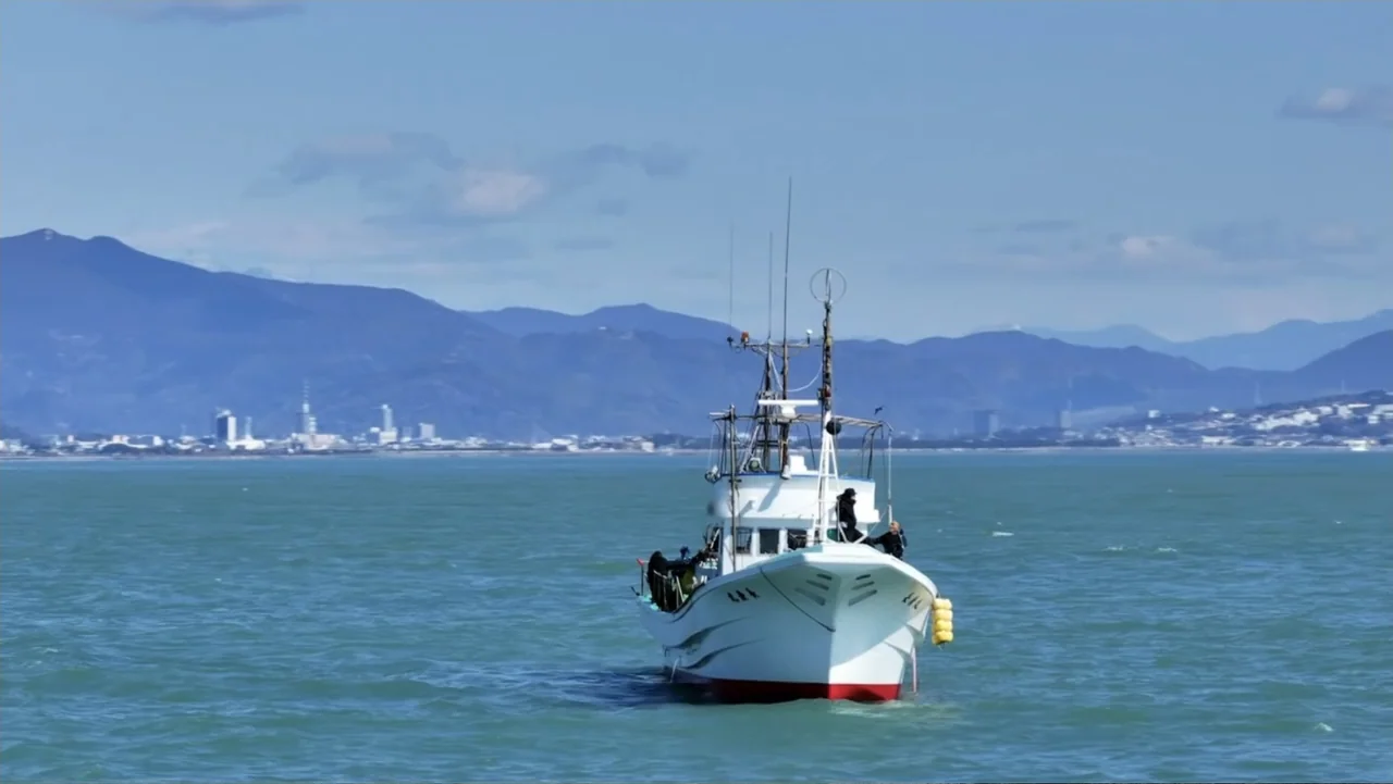 Alien Sharks: Ghosts of Japan (2024) movie still: wide — A white fishing boat is positioned in calm coastal waters under a clear sky. Distant…
