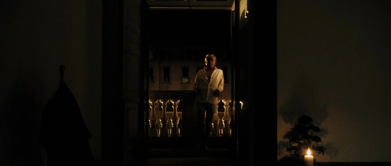 The Great Beauty (2013) movie still: fullbody — A dimly lit room overlooks a dark stone balcony at night. An elderly man in white clothes stands on…