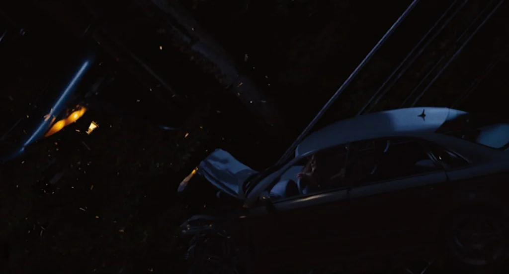 Louder Than Bombs (2015) movie still: wide — A dark, angled scene shows a crashed vehicle off the side of a road. A person sits in the driver's seat…