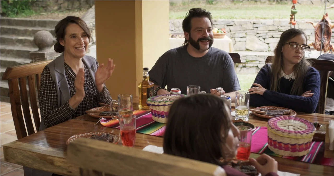 Bendita Suegra (2023) movie still: medium — A group gathers around a large wooden table on a covered stone terrace patio. Three adults and one…