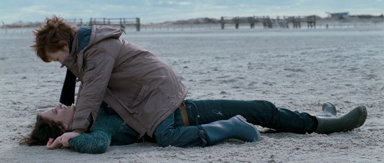 Womb (2010) movie still: fullbody — A wide, flat sand beach under an overcast sky stretches toward a distant pier. One person kneels in the sand…