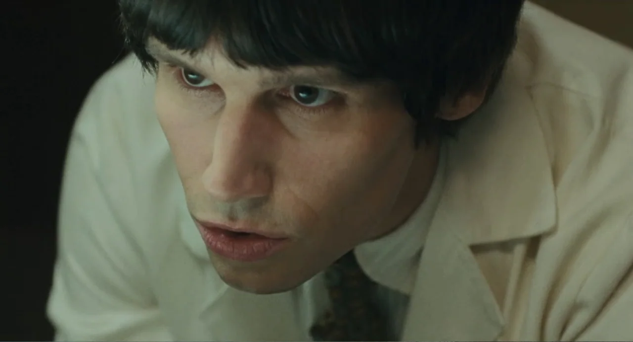 Call Jane (2022) movie still: closeup — A man stands inside a room with cream walls wearing a light button-down shirt. The man gazes upward with a…