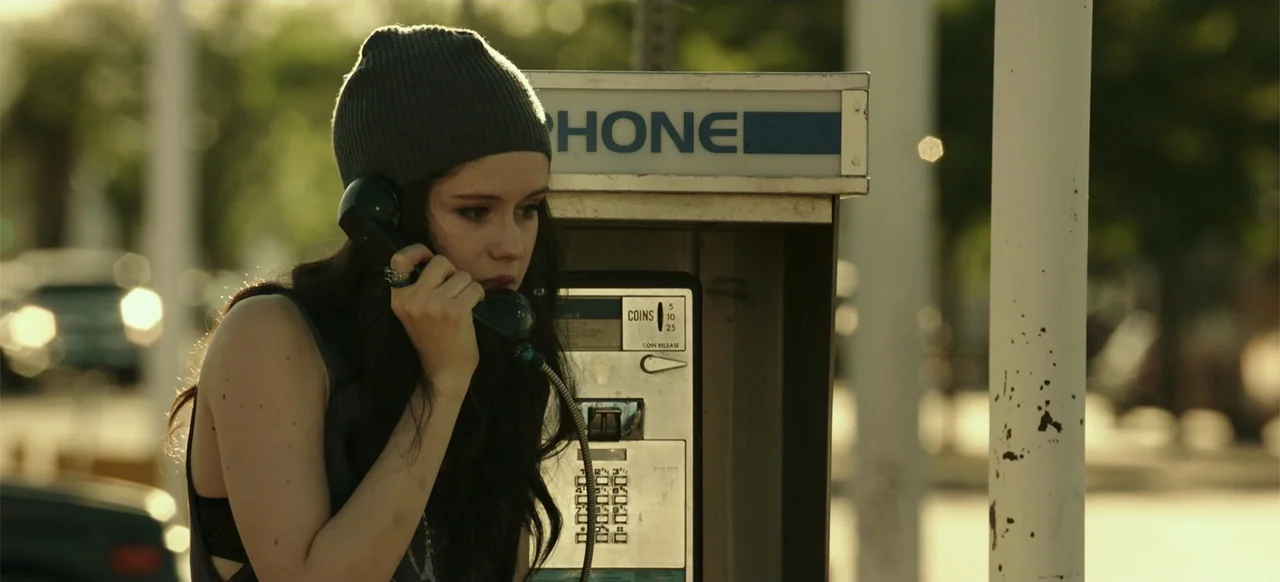 Blood Father (2016) movie still: medium — A public outdoor payphone booth is positioned on a city street at sunset. A young woman in a beanie uses…