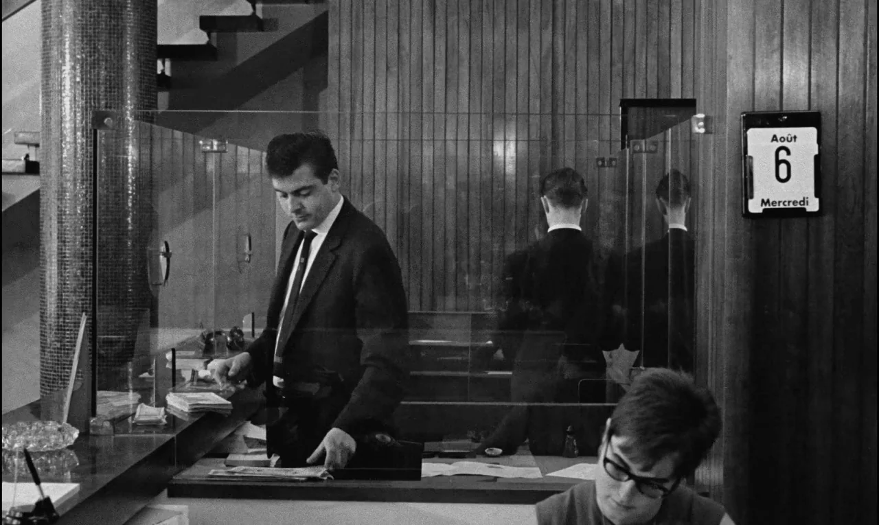 Bay of Angels (1963) movie still: medium — A teller stands behind a clear glass divider in a wood-paneled bank branch. He organizes currency on the…