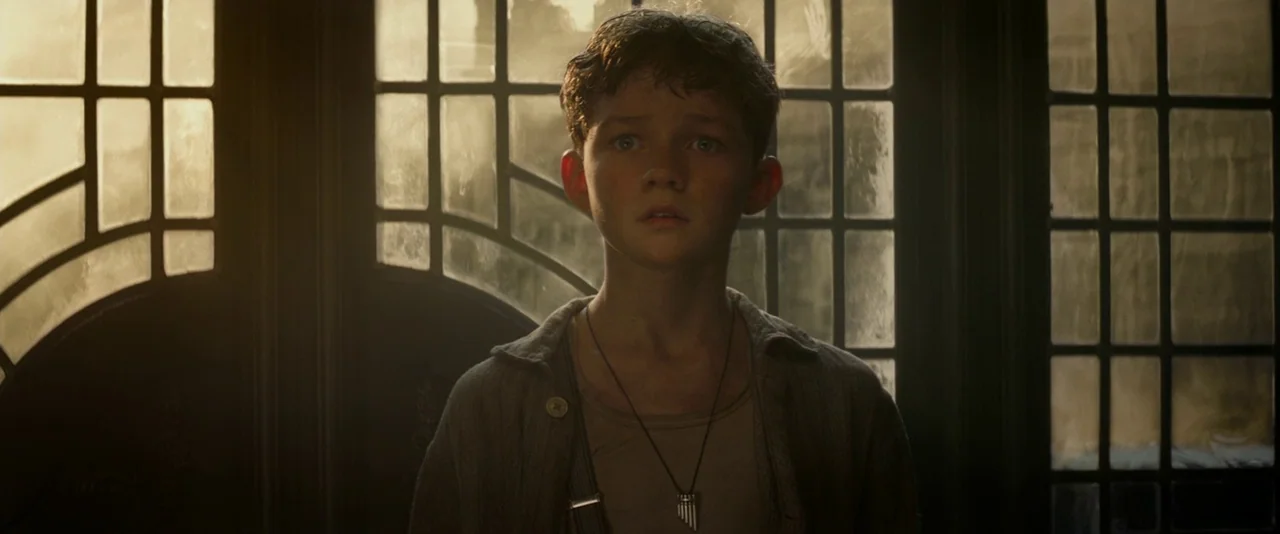 Pan (2015) movie still: medium — A young boy stands before a large, multi-paned glass door with dark metal frames. He gazes forward with a neutral…