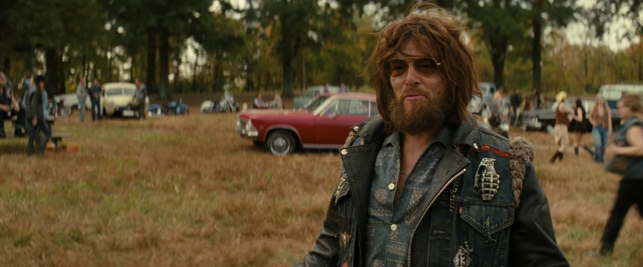 The Bikeriders (2023) movie still: medium — A grassy outdoor field populated with vintage cars and several people in the distance. A man with a…