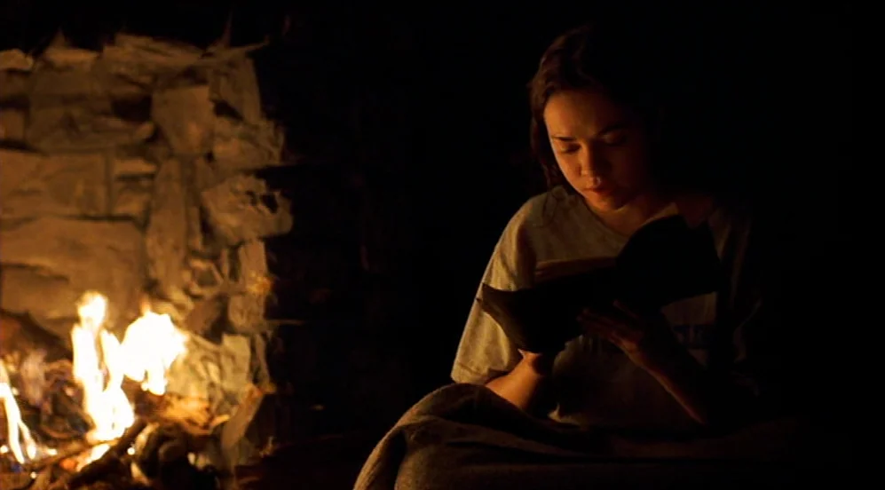 Limbo (1999) movie still: medium — A dimly lit space features a stone fireplace with active flames. A young woman reads a book while wrapped in a…