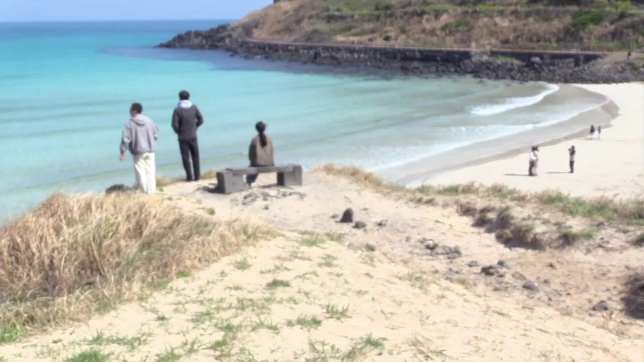 In Water (2023) movie still: wide — A sandy cliff edge overlooks a turquoise bay with a beach below. Three people stand and sit on the cliff, while…