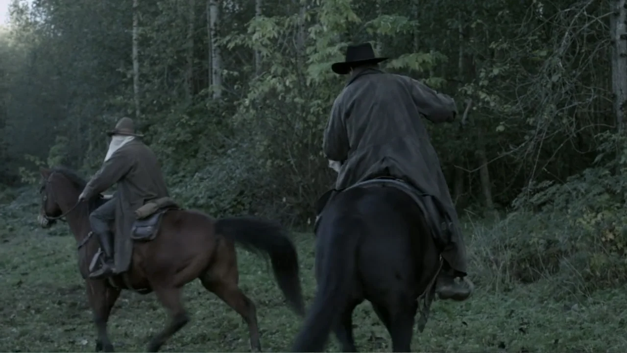 Dawn Rider (2012) movie still: fullbody — Two men on horseback travel through a dense forest with tall pine trees. The riders wear long dark coats…