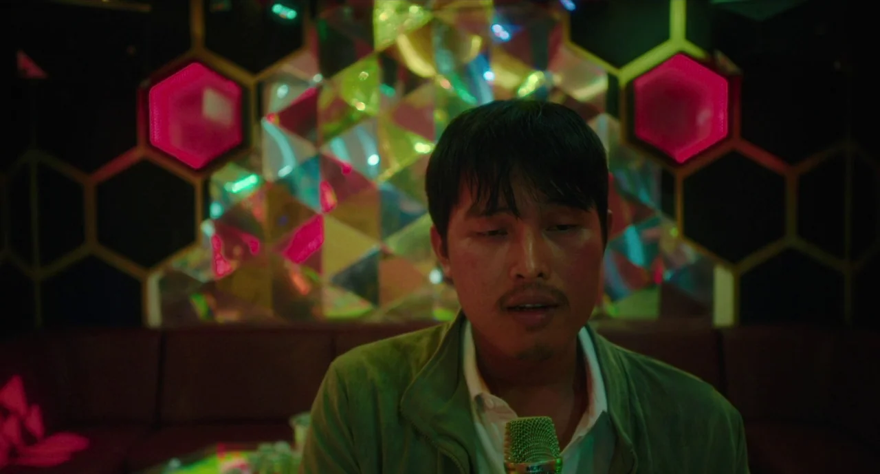 Inside The Yellow Cocoon Shell (2023) movie still: medium — A dimly lit karaoke room features a decorative wall panel made of geometric mirrors. A…
