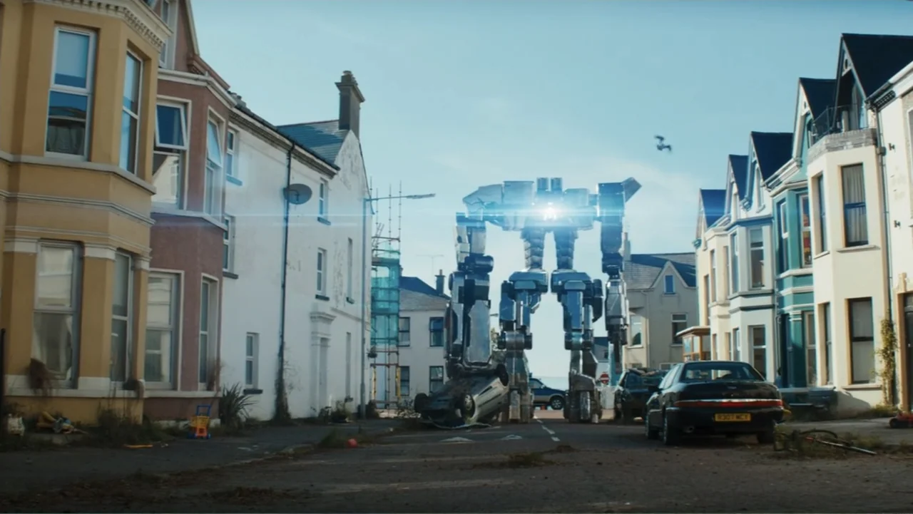 Robot Overlords (2015) movie still: wide — A residential street is lined with multi-story houses under a clear blue sky. A tall mechanical robot…
